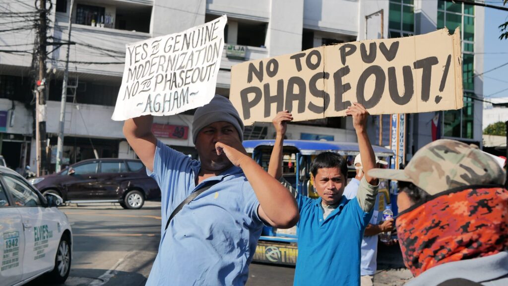 Two jeepney drivers in a picketline during a transport strike hold a sign that says "Yes to genuine modernization, no to phaseout" and "No to PUV Phaseout"